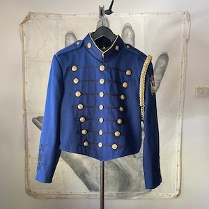 K&ouml;nnte beinhalten: Eine blaue Jacke im Milit&auml;rstil mit goldenen Kn&ouml;pfen und schwarzen Verzierungen. Die Jacke hat einen Stehkragen und lange &Auml;rmel. Am rechten &Auml;rmel befindet sich eine goldene Litzenverzierung.