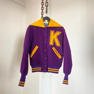 Può includere: Giacca da baseball viola e oro con una grande lettera "K" sul davanti. La giacca ha un colletto giallo e strisce gialle sulle maniche e sul fondo.