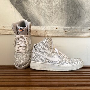 May include: White high-top sneakers with a white Nike swoosh and the words "Just Do It" printed all over the shoe. The words "Nike Air" are printed on the tongue of the shoe.