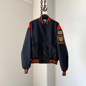 Puede incluir: Chaqueta bomber negra de satén con ribetes de rayas naranja y blancas. La chaqueta tiene un parche en la manga izquierda con el texto "Walker Rogers" y "Post 082".