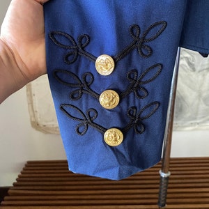 K&ouml;nnte beinhalten: Ein marineblauer Blazer mit schwarzen Stickereien und goldenen Kn&ouml;pfen. Die Kn&ouml;pfe haben ein erhabenes Design.