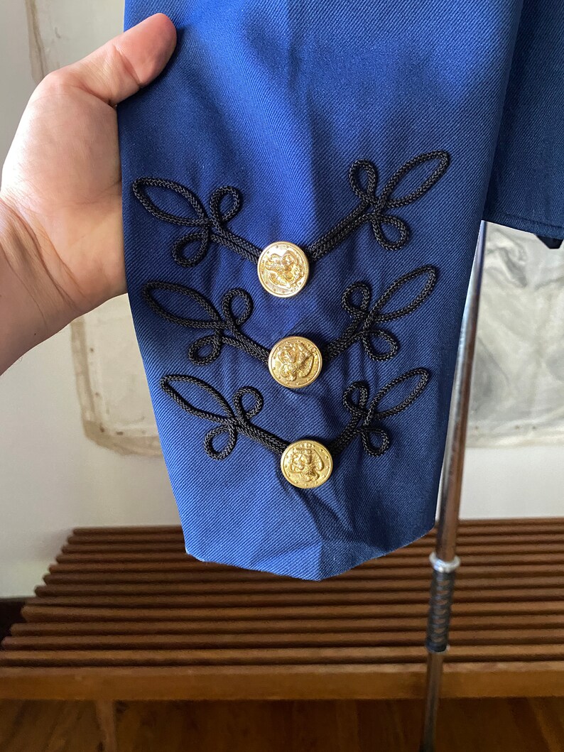 K&ouml;nnte beinhalten: Ein marineblauer Blazer mit schwarzen Stickereien und goldenen Kn&ouml;pfen. Die Kn&ouml;pfe haben ein erhabenes Design.