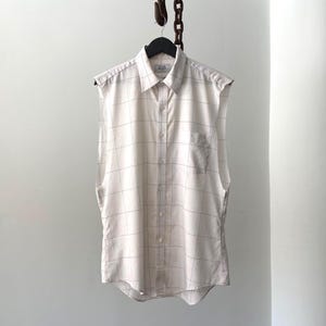 Op de afbeelding: Een mouwloos crème overhemd met een rasterpatroon, hangend aan een zwarte hanger. Het overhemd heeft een kraag, een borstzak en is aan de voorkant dichtgeknoopt. De lichte stof is neutraal van kleur.