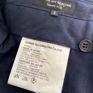 K&ouml;nnte beinhalten: Ein dunkelblaues Kleidungsst&uuml;ck-Etikett mit wei&szlig;em Text, der "COMME des GAR&Ccedil;ONS Co., Ltd." lautet und die Stoffzusammensetzung und Pflegehinweise angibt. Das Etikett enth&auml;lt auch den Text "MADE IN JAPAN" und "FABRIQUE AU JAPON".