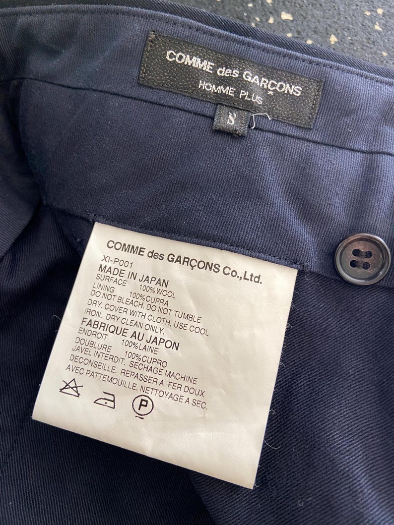 K&ouml;nnte beinhalten: Ein dunkelblaues Kleidungsst&uuml;ck-Etikett mit wei&szlig;em Text, der "COMME des GAR&Ccedil;ONS Co., Ltd." lautet und die Stoffzusammensetzung und Pflegehinweise angibt. Das Etikett enth&auml;lt auch den Text "MADE IN JAPAN" und "FABRIQUE AU JAPON".