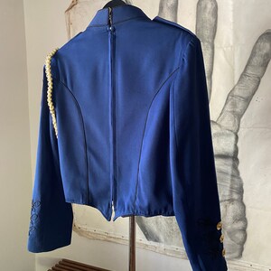 K&ouml;nnte beinhalten: Ein marineblauer Blazer mit einem goldenen Kettendetail an der Schulter, schwarzen Stickereien an den &Auml;rmeln und goldenen Kn&ouml;pfen. Der Blazer hat einen Rei&szlig;verschluss auf der R&uuml;ckseite.