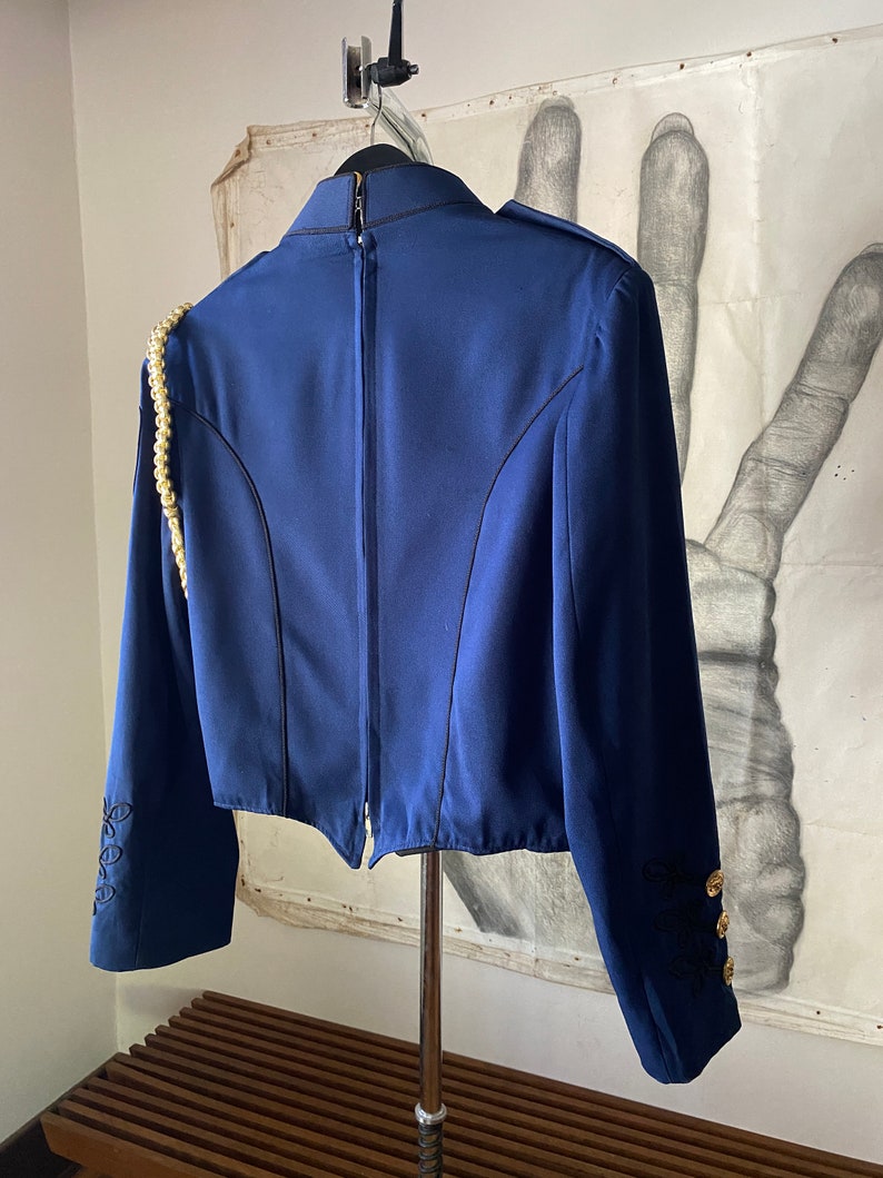 K&ouml;nnte beinhalten: Ein marineblauer Blazer mit einem goldenen Kettendetail an der Schulter, schwarzen Stickereien an den &Auml;rmeln und goldenen Kn&ouml;pfen. Der Blazer hat einen Rei&szlig;verschluss auf der R&uuml;ckseite.