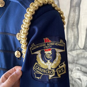 K&ouml;nnte beinhalten: Eine marineblaue Jacke mit goldener Litzenbesatz und einem Aufn&auml;her auf dem linken &Auml;rmel. Der Aufn&auml;her ist schwarz mit goldener Stickerei und tr&auml;gt die Aufschrift "Knowledge Achievement Unity" und "Carencro HS".
