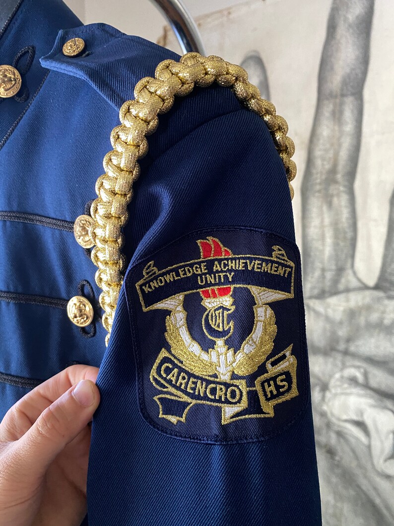 K&ouml;nnte beinhalten: Eine marineblaue Jacke mit goldener Litzenbesatz und einem Aufn&auml;her auf dem linken &Auml;rmel. Der Aufn&auml;her ist schwarz mit goldener Stickerei und tr&auml;gt die Aufschrift "Knowledge Achievement Unity" und "Carencro HS".