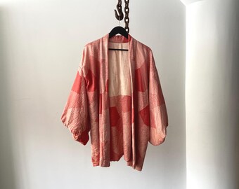 May include: A coral and peach-coloured kimono-style jacket with a geometric pattern. The jacket is hanging on a black hanger suspended from a chain. The sleeves are wide and the fabric appears textured.