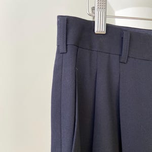 K&ouml;nnte beinhalten: Eine dunkelblaue, gefaltete Hose, die an einem Kleiderb&uuml;gel h&auml;ngt. Die Hose hat eine gerade Bein und einen klassischen Stil.