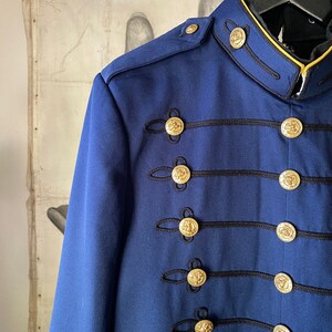 K&ouml;nnte beinhalten: Eine marineblaue Jacke mit goldenen Kn&ouml;pfen und schwarzem Besatz. Die Jacke hat einen Stehkragen und einen einreihigen Verschluss.