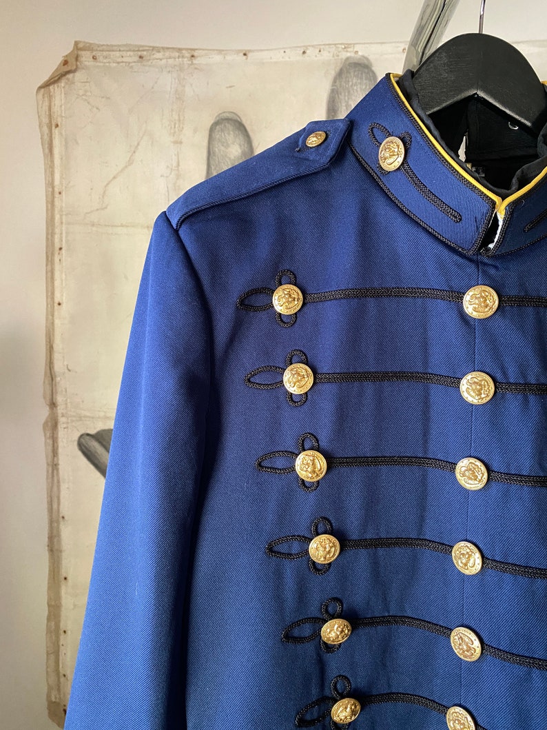 K&ouml;nnte beinhalten: Eine marineblaue Jacke mit goldenen Kn&ouml;pfen und schwarzem Besatz. Die Jacke hat einen Stehkragen und einen einreihigen Verschluss.