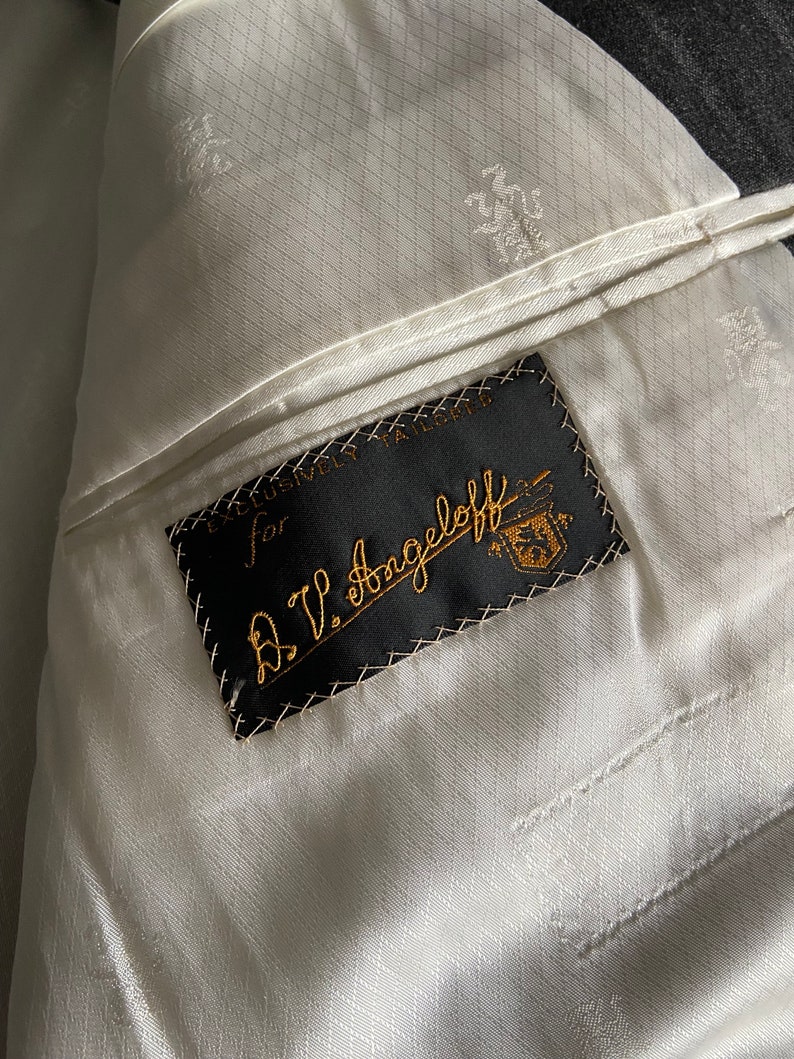 May include: A black label sewn into a white fabric lining with the text "Exclusively Tailored for A.V. Angell off" in white cursive lettering.