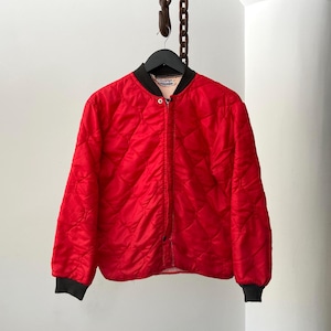 May include: A bright red quilted jacket featuring a diamond-patterned stitch, a black ribbed collar, and black ribbed cuffs. The jacket has a central zipper closure and a light-colored interior lining visible at the neckline, hanging on a black hanger.