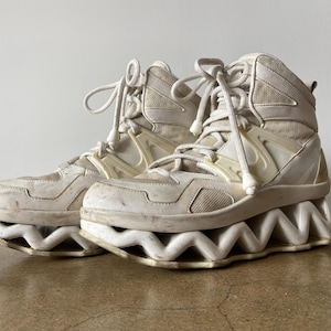 Puede incluir: Zapatillas altas de color blanco roto con un diseño de suela ondulada único. Las zapatillas combinan malla y cuero sintético, con cierre de cordones. Tienen un diseño futurista distintivo, con una suela en zigzag.