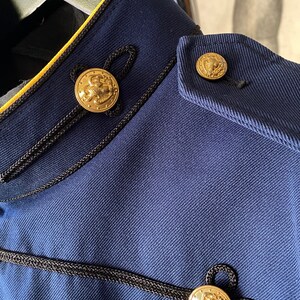 K&ouml;nnte beinhalten: Nahaufnahme einer marineblauen Jacke mit schwarzem und gelbem Besatz. Die Jacke hat drei goldene Kn&ouml;pfe mit einem erhabenen Design.