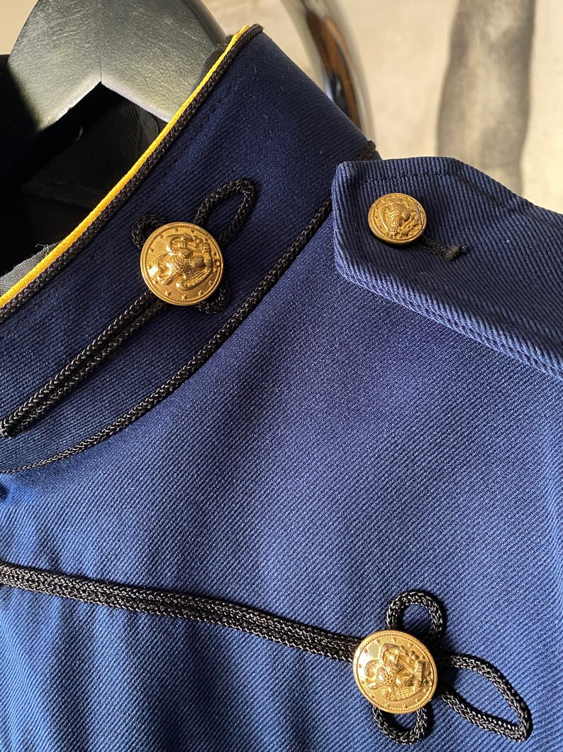 K&ouml;nnte beinhalten: Nahaufnahme einer marineblauen Jacke mit schwarzem und gelbem Besatz. Die Jacke hat drei goldene Kn&ouml;pfe mit einem erhabenen Design.