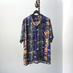 May include: A short-sleeved button-down shirt with a navy blue background and a tropical print featuring palm trees, flowers, and other island imagery. The shirt is hanging on a black hanger.