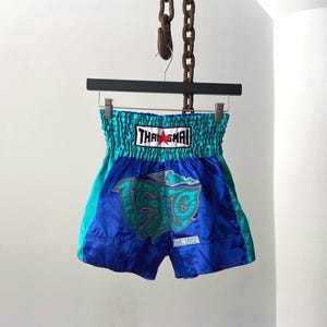 May include: Blue and teal Muay Thai shorts with a teal waistband and a large graphic on the front. The shorts have the text "THAI SMAI" on the waistband and "est. 1972" on the front.