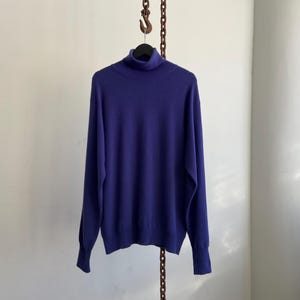 May include: A solid purple turtleneck sweater hangs from a black hanger. The long-sleeved sweater is made of a soft material and has ribbed cuffs. The background is a plain white wall.