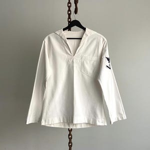 Peut inclure: Une chemise de marin blanche à manches longues, avec un col en V et une petite poche poitrine. La chemise a une coupe ample et un emblème bleu marine sur la manche gauche. Elle est suspendue à un cintre noir.