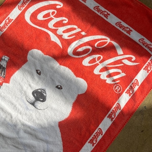 May include: Red beach towel featuring a white polar bear and the Coca-Cola logo in white lettering. The towel has a white border with the Coca-Cola logo repeated around the edge.