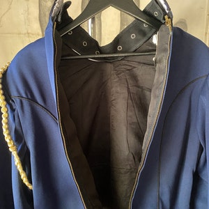 K&ouml;nnte beinhalten: Eine marineblaue Jacke mit einem goldenen Kettendetail an der linken Schulter. Die Jacke hat einen Rei&szlig;verschluss und ein schwarzes Futter. Der Kragen hat ein schwarzes Band mit Metall&ouml;sen.