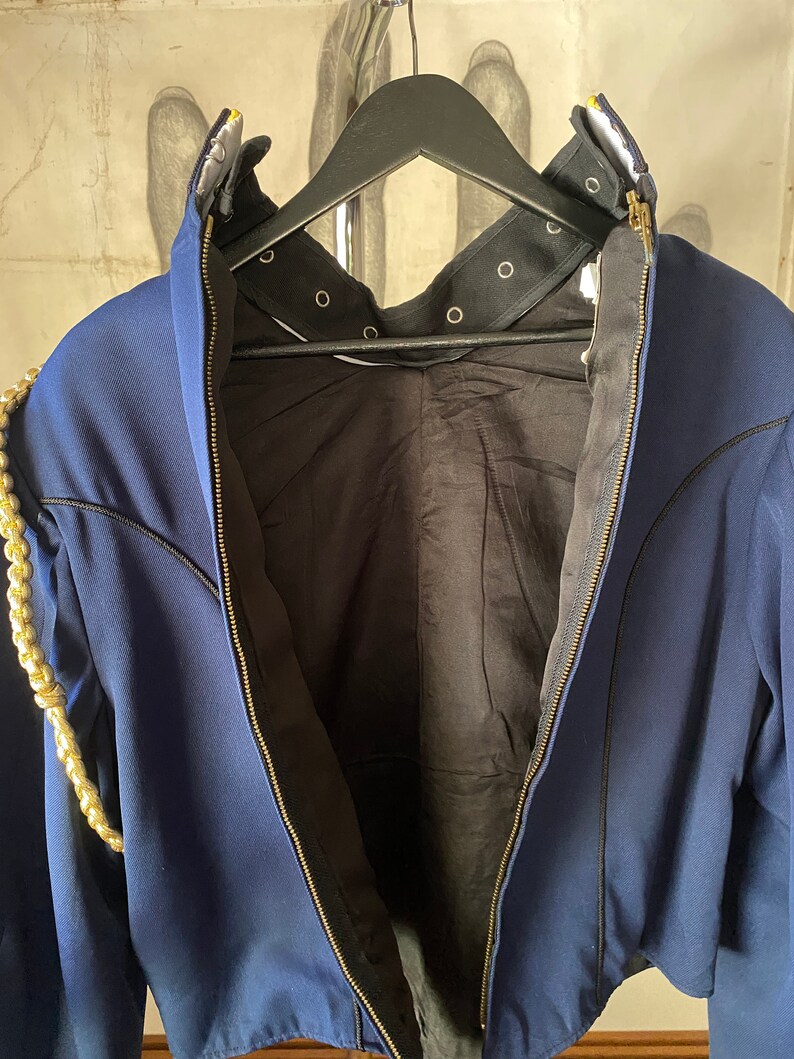 K&ouml;nnte beinhalten: Eine marineblaue Jacke mit einem goldenen Kettendetail an der linken Schulter. Die Jacke hat einen Rei&szlig;verschluss und ein schwarzes Futter. Der Kragen hat ein schwarzes Band mit Metall&ouml;sen.