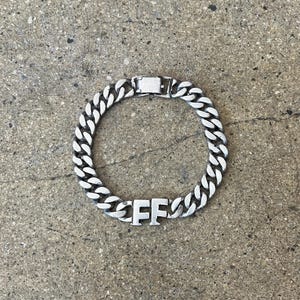 Puede incluir: Pulsera de cadena de eslabones de color plateado con cierre rectangular. La pulsera presenta las letras "FF" en el centro. La joya se exhibe sobre una superficie gris texturizada.