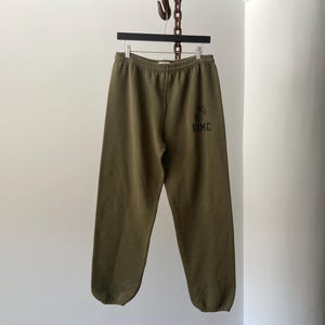 May include: Olive green sweatpants with a drawstring waist and elastic cuffs. The sweatpants have a black embroidered graphic of a military symbol and the text "USMC" on the left leg.