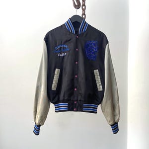 Puede incluir: Chaqueta universitaria negra y gris con detalles de rayas azules y blancas. Presenta el texto "Carson High Colts" y un gran parche azul con la letra "C". La chaqueta tiene mangas grises y botones rosas.