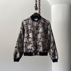 May include: A black and gold bomber jacket with a floral pattern. The jacket has a zipper closure and two pockets.