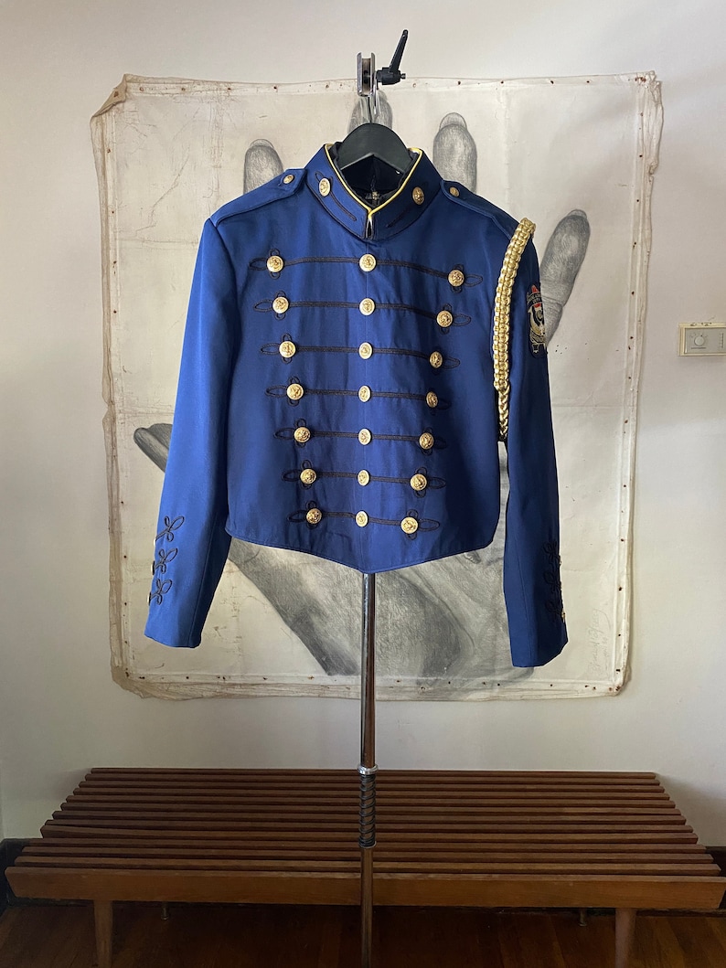 K&ouml;nnte beinhalten: Eine blaue Jacke im Milit&auml;rstil mit goldenen Kn&ouml;pfen und schwarzen Verzierungen. Die Jacke hat einen Stehkragen und lange &Auml;rmel. Am rechten &Auml;rmel befindet sich eine goldene Litzenverzierung.