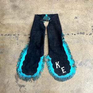 Puede incluir: Chaps de ante negro con bordes festoneados y flecos turquesa. Los chaps tienen un cierre de hebilla en la cintura y las letras "K E" en blanco en una pierna. Ropa del oeste.