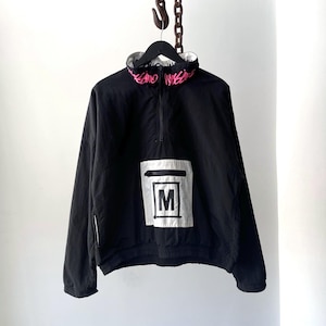 May include: Black anorak jacket with a white and silver square patch featuring the letter "M". The collar has pink text. The jacket has a half-zip closure and a white lining.