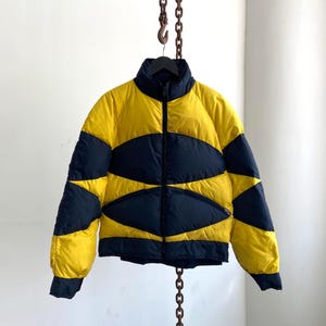 May include: A yellow and navy blue puffer jacket with a high collar and a full-length zipper. The jacket features a unique design with curved navy blue panels across the chest and sleeves. The jacket is hanging from a metal chain.