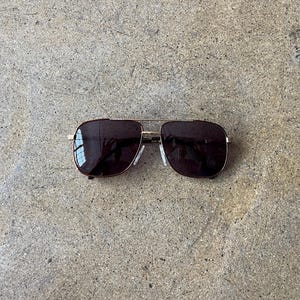 May include: Sunglasses with a gold-colored frame and dark lenses. The sunglasses have a classic aviator style with a double bridge and a rectangular shape. The lenses are a dark brown color, and the frame has a shiny finish.