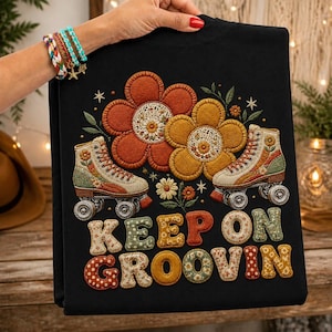 May include: A black t-shirt featuring an embroidered design of two roller skates and two large flowers. The text "KEEP ON GROOVIN" is displayed in large, patterned, multi-colored letters. The design includes small stars and floral accents.