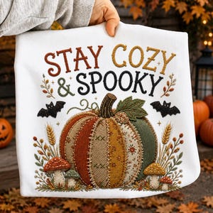 以下が含まれることがあります： STAY COZY & SPOOKYという文字が刺繍された白い布製品。パッチワーク風の大きなカボチャ、2匹の黒いコウモリ、小麦の茎、そしてオレンジ、緑、茶色の小さなキノコが描かれたデザインが特徴です。