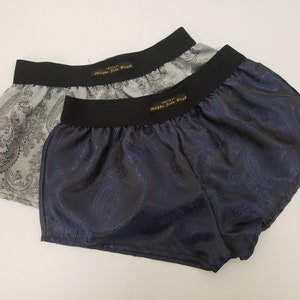 May include: Two pairs of silk boxer shorts with a paisley pattern. The top pair is light grey with a black waistband and the bottom pair is dark blue with a black waistband. Both pairs have the text "Philippe Jude Wright" printed in gold on the waistband.