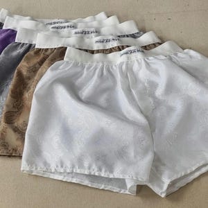 Peut inclure: Une collection de shorts de boxe en satin de différentes couleurs, dont blanc, marron, gris, violet et bleu. Les shorts ont une ceinture blanche et un motif floral subtil. Les shorts sont disposés en éventail.