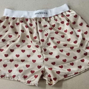 Op de afbeelding: Crème satijnen boxershort met een patroon van rode harten. De short heeft een witte tailleband met tekst. Een casual en comfortabel kledingstuk.