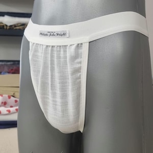 Könnte beinhalten: Weißer Jockstrap aus leichtem, strukturiertem Stoff. Der Bund und die Kanten sind glatt, mit einem kleinen Etikett mit der Aufschrift "CREATED BY Philippe John Wright". Der Jockstrap wird auf einer grauen Schaufensterpuppe präsentiert.