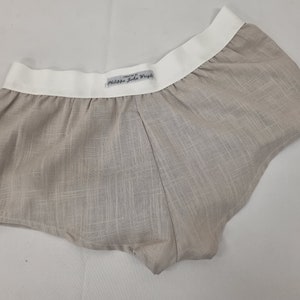 Può includere: Shorts di lino beige con elastico in vita bianco. La cintura ha un'etichetta con la scritta "Designed by Philippe John Wright".
