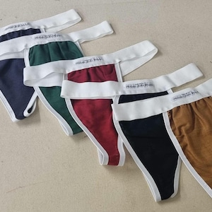 Op de afbeelding: Een verzameling sportondersteuners in verschillende kleuren: marineblauw, groen, bordeauxrood, zwart en bruin. Elke ondersteuner heeft een witte tailleband en bies, met tekst op de tailleband.