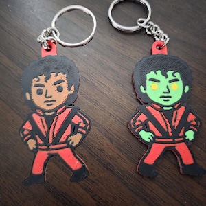 May include: Two keychains featuring cartoon figures in red and black outfits. One figure has brown skin and the other has green skin. Both have black hair and are attached to silver keyrings.