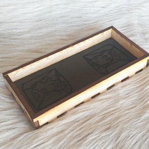 Puede incluir: Una bandeja de dados de madera con un forro de fieltro marrón oscuro. La bandeja tiene forma rectangular y presenta dos dados grabados con un diseño negro.