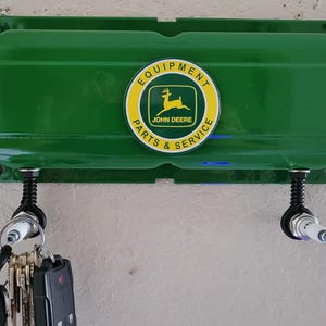 May include: Green metal wall-mounted key holder with a John Deere logo. The logo is yellow and green and says "Equipment Parts & Service". There are two key hooks on the holder.