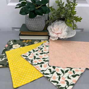 May include: Four different fabric placemats with various patterns. One placemat is green with white flowers, one is yellow with white polka dots, one is pink with white stripes, and one is white with green leaves and pink flowers.
